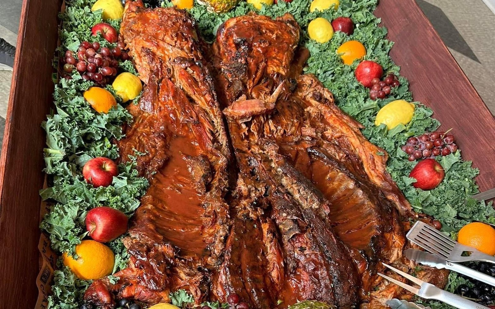 A full catering tray of smoked ribs and pulled pork, garnished with fresh fruit and greens