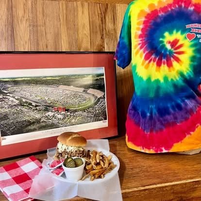 A plate of fried pork chop, mac and cheese, and fries on a checkered tablecloth, with a framed Darlington Raceway print in the background
